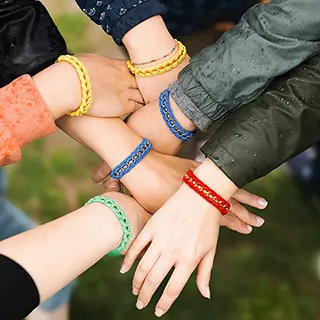 Un cercle d'amies montre leurs poignets liés par des bracelets colorés de l'amitié, symbolisant les liens et la diversité au sein du groupe, avec des nuances de rouge, bleu, jaune et vert, sur un fond naturel.