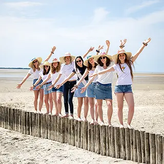 Les bras levés et mains jointes, un groupe d'amies en chapeaux de paille et tenues estivales crée une chaîne humaine joyeuse sur une plage de sable, symbolisant l'unité et l'amitié.