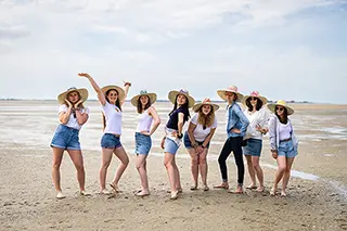 Sept femmes rayonnantes, coiffées de chapeaux de paille et vêtues de tenues décontractées, posent avec assurance sur une plage, transmettant un esprit d'aventure et de liberté.