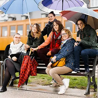 Un moment de complicité capturé entre six amies assises sur un banc à Saint-Valery-sur-Somme, avec des parapluies ouverts, partageant des sourires et des conversations en attendant le train en arrière-plan.
