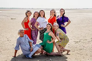 Un groupe de huit femmes, vêtues de robes colorées, prend la pose sur une plage de sable, incarnant l'élégance et la joie de vivre sous un ciel lumineux.