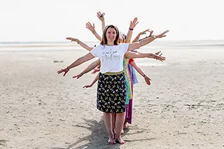 Une femme au centre, les bras étendus, est entourée par des amies positionnées de manière à former une étoile humaine, un jeu créatif et joyeux sur une plage aux tons clairs.