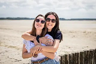 Deux amies s'étreignent tendrement sur la plage, leurs lunettes de soleil reflétant le ciel clair, un moment de complicité et de bonheur partagé en plein air.