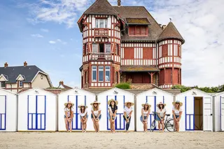 Devant une maison normande traditionnelle, un groupe de femmes en robes et chapeaux de paille pose aligné devant des cabines de plage blanches et bleues, évoquant le charme intemporel du bord de mer.
