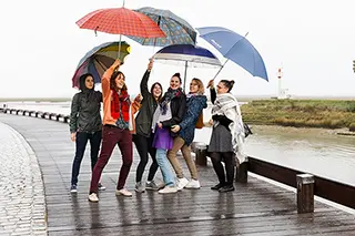 Un groupe de sept personnes joyeuses, armées de parapluies colorés, célèbre un moment ensemble sur un chemin pavé le long de la côte à Saint-Valery-sur-Somme. Malgré un ciel gris et une atmosphère pluvieuse, l'ambiance est chaleureuse et festive.