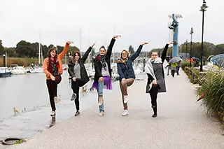 Cinq femmes expriment leur joie en levant une jambe dans un élan de danse spontané, sur le chemin du port de Saint-Valery-sur-Somme, évoquant une célébration vivante et décontractée en dépit du ciel couvert.