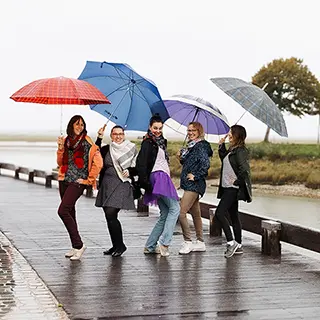 Un groupe de six femmes partage un moment joyeux sous des parapluies colorés en marchant sur une jetée pavée, malgré le temps gris et pluvieux.