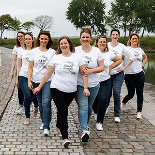 Un groupe de huit femmes marchant côte à côte sur le quai pavé, toutes vêtues de t-shirts blancs imprimés et jeans, dégageant une ambiance détendue et heureuse.
