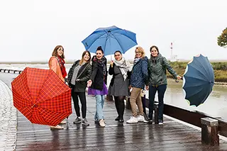 Six femmes se promenant sur un quai par un jour de pluie, souriantes et animées, utilisant leurs parapluies pour ajouter une touche de couleur à la scène grisâtre.