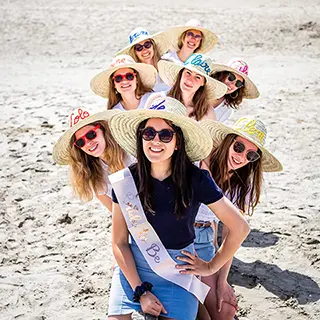 Un groupe souriant de femmes formant une pyramide humaine sur une plage ensoleillée, toutes coiffées de chapeaux de paille décorés, symbolisant l'amusement et la cohésion du groupe.