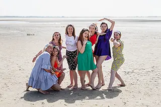 Une scène joviale de femmes en robes colorées et une femme en position accroupie, toutes partageant un éclat de rire sur la plage, capturant une ambiance détendue et heureuse.