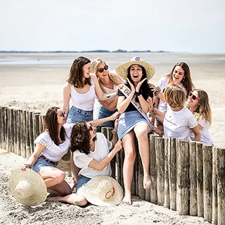 La bonne humeur est palpable parmi ce groupe d'amies assises sur une dune, s'amusant et riant ensemble, complétées par leurs chapeaux de paille personnalisés, sur fond de ciel bleu et sable fin.