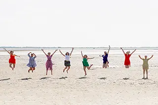 Huit femmes en robes colorées s'élèvent en un saut de joie sur une vaste plage, les bras levés vers le ciel clair, capturant un moment de bonheur partagé et d'insouciance.