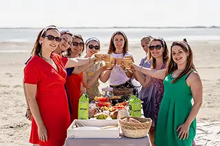 Un groupe de femmes célébrant avec un toast de sandwichs et de boissons sur une nappe posée sur le sable de la plage, partageant un pique-nique convivial sous le soleil.
