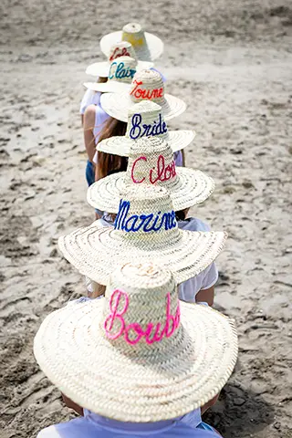 Vue arrière d'une file indienne de femmes portant des chapeaux de paille personnalisés avec leurs noms écrits en lettres colorées, debout sur une plage, créant une perspective amusante et originale.