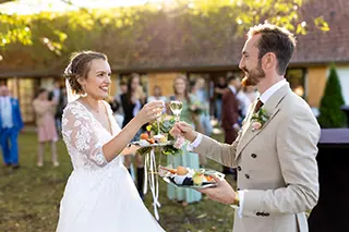 Mariés portant un toast avec des coupes de champagne, avec des invités en arrière-plan lors d'une réception en plein air.