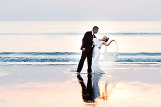 Mariés partageant un moment intime sur une plage au coucher du soleil, se reflétant sur l'eau.