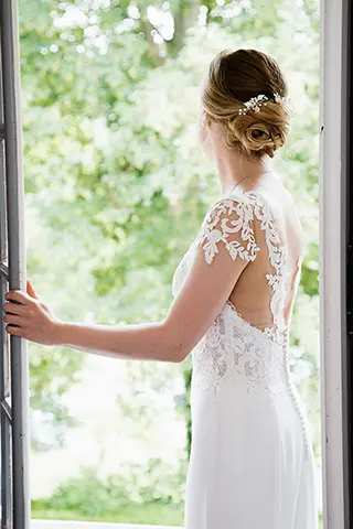 Mariée avec un regard lointain devant une fenêtre, dos nu de la robe