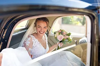 Mariée souriante en robe de dentelle tenant un bouquet de fleurs à l'intérieur d'une voiture classique.