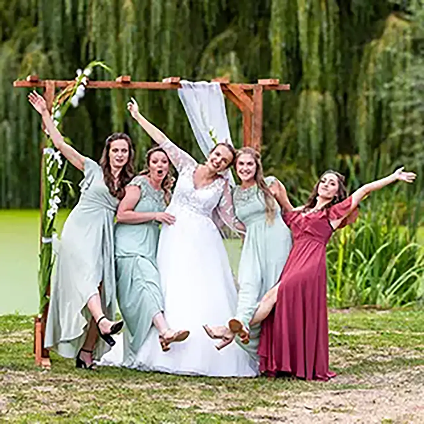 Mariée joyeuse avec ses demoiselles d'honneur posant de manière espiègle sous un arche de mariage dans un jardin.