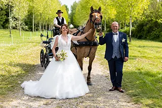 Mariée souriante tenant la bride d'un cheval à côté du marié et d'une calèche avec cocher en arrière-plan.