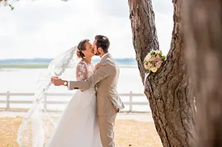 Un couple de jeunes mariés s'embrasse sous un pin avec un plan d'eau en arrière plan