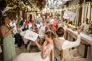 Mariés jouant à un jeu pendant la soirée de mariage, tenant des pancartes 'LUI' pour répondre à des questions