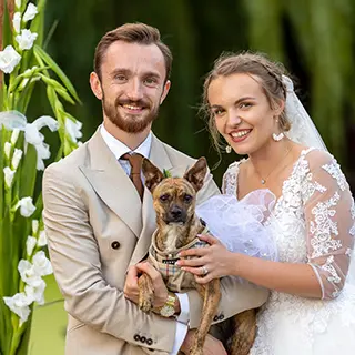 Mariés souriants tenant leur chien entre eux, avec des fleurs blanches et un feuillage vert en arrière-plan.