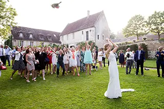 La mariée lançant son bouquet vers un groupe de femmes invitées dans l'attente, lors d'un mariage en extérieur avec une maison en pierre en arrière-plan.