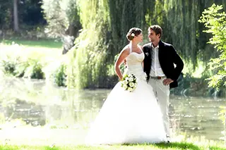 Mariés posant dans un parc verdoyant avec un étang en arrière-plan, échangeant un regard complice.