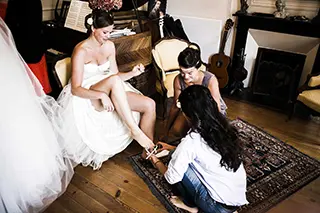 Une mariée assise est aidée par deux femmes à mettre ses chaussures dans un salon élégamment décoré.