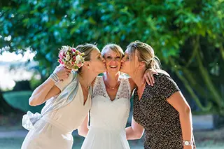 La mariée reçoit des baisers sur les joues de deux invitées dans un cadre extérieur verdoyant.