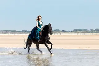 Une femme souriante en robe sur un cheval noir au galop sur la plage