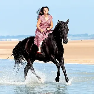 Une femme en robe légère sur un cheval noir au galop sur la plage