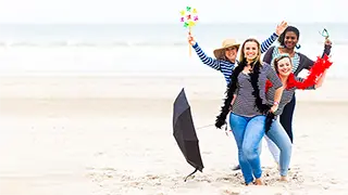 Groupe de quatre amis souriants marchant sur une plage, tenant des parapluies et un moulin à vent, signifiant la joie et l'amitié.