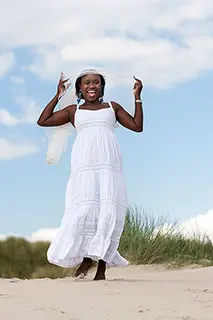 Femme en robe blanche souriante et dansant sur une dune avec un ciel bleu en arrière-plan.