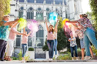 Groupe de personnes célébrant avec des poudres colorés devant une église gothique, exprimant la joie et l'éclat dans une atmosphère festive.