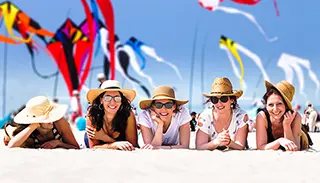 Groupe de femmes souriantes allongées sur le sable avec des chapeaux et des lunettes de soleil, avec des cerfs-volants colorés volant dans le ciel bleu en arrière-plan.
