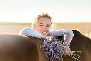 Jeune fille souriante adossée au dos d'un cheval tenant un bouquet de fleurs violettes dans la main
