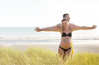 Couple souriant sur la plage, l'homme derrière la femme enceinte, les bras ouverts dans un geste d'envol