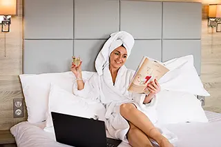 Femme souriante en peignoir et avec une serviette sur la tête, tenant un verre de vin et un livre dans une chambre d'hôtel