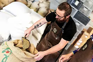 Un homme en tablier maron prends prends une pelleté de café en grain dans un sac en toile de jute