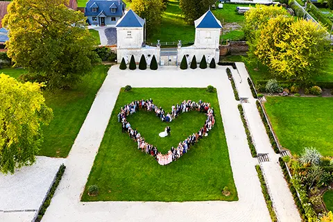 Vue aérienne d'un rassemblement formant un cœur sur la pelouse verte des jardins d'un château, entourée d'allées et de bâtiments élégants, lors d'un événement festif