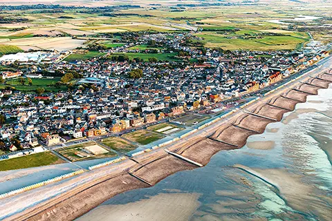 Vue aérienne d'une ville côtière avec des plages, des cabines de bain alignées, et des champs verdoyants en arrière-plan